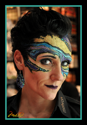 Monique Lily in a gold, blue, and purple mask painted by Mark Reid